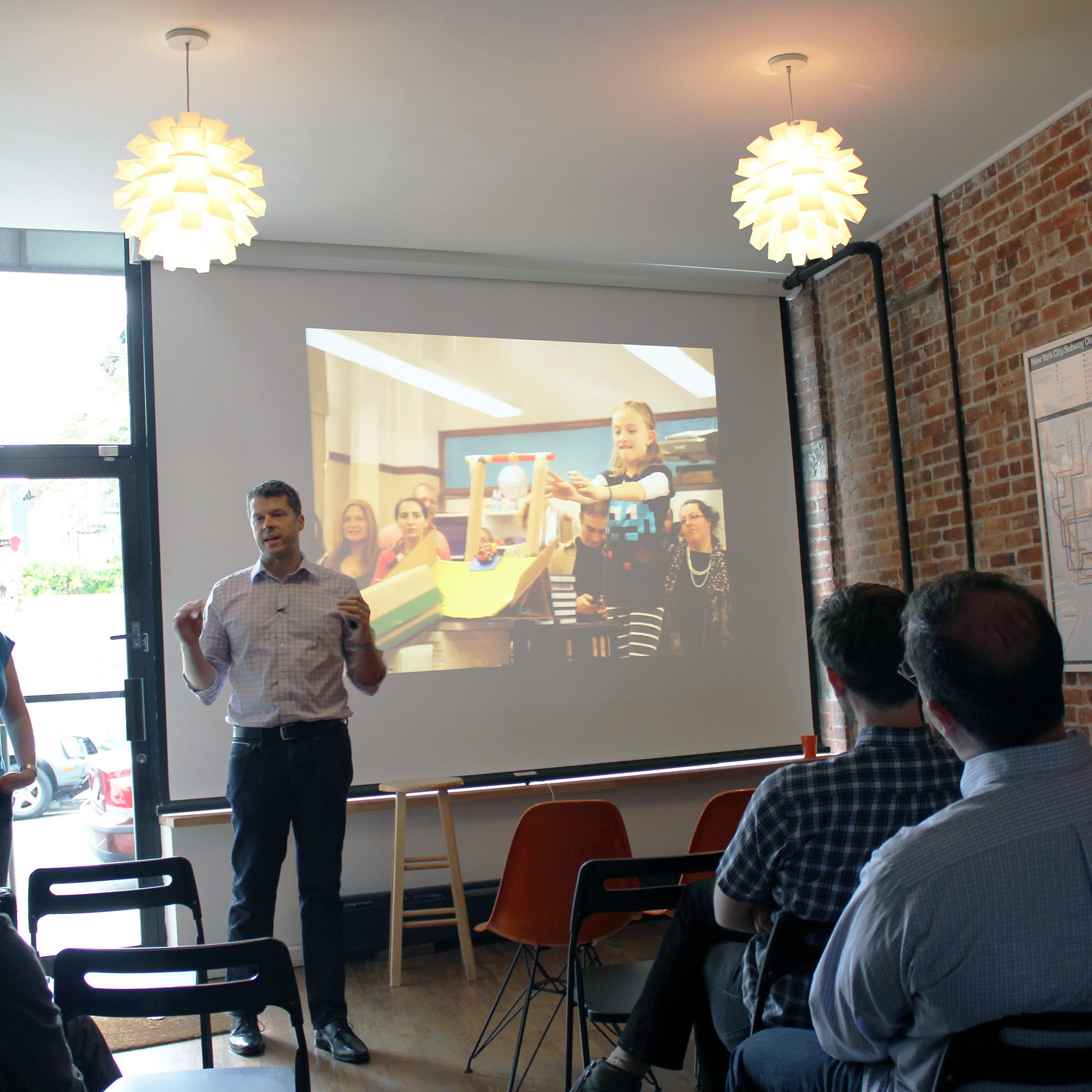 A group of people sit facing a man and woman who are presenting in front of a screen in a modern room with brick walls. The screen displays a photo of children engaged in an activity. The presenters stand near a large window, and pendant lights hang from the ceiling.
