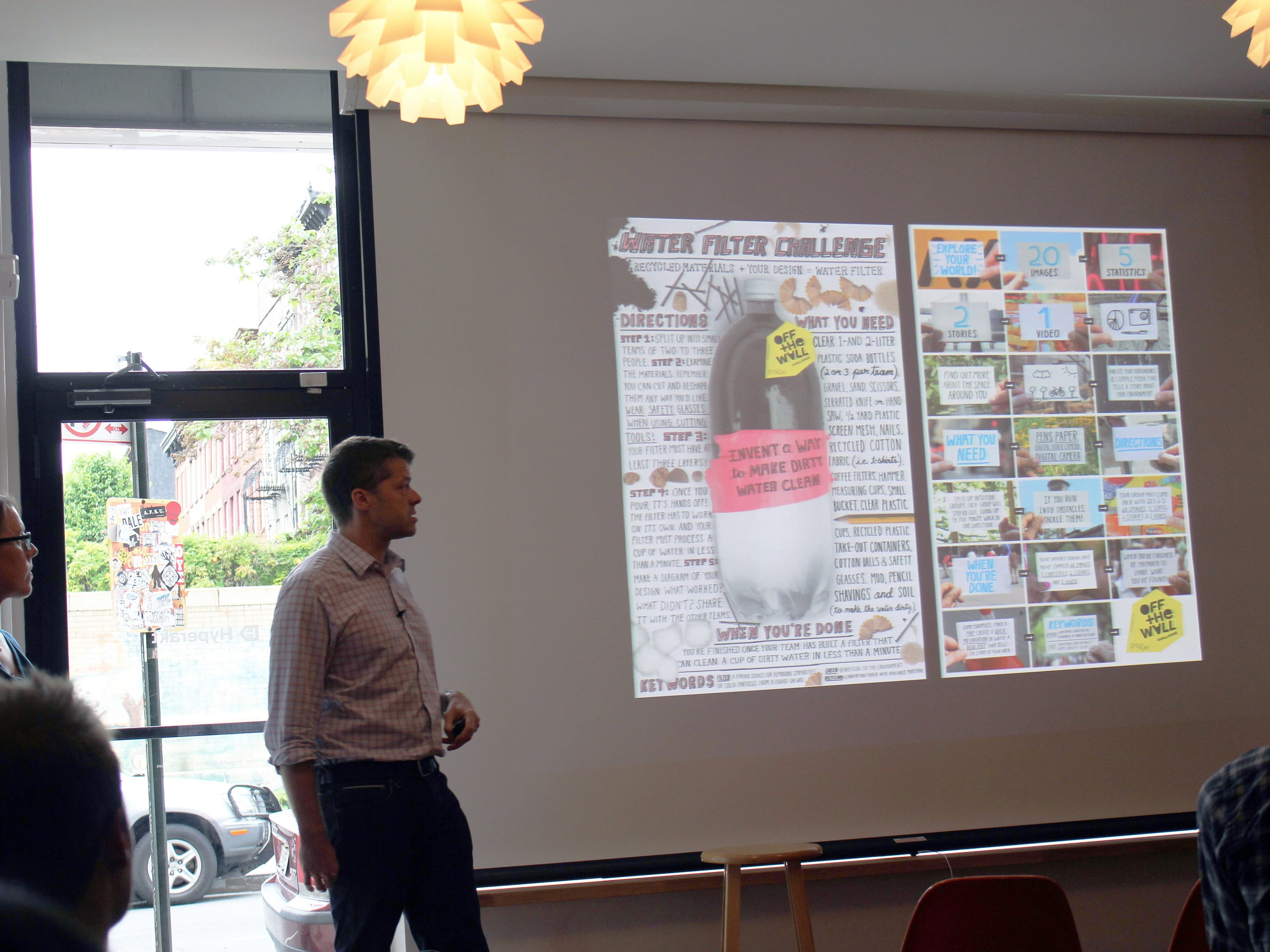 A man and a woman give a presentation in a room with a large screen showing colorful graphics and text about the "Butterfly Cup Challenge". The room has large windows and modern lighting fixtures. Several people are seated, facing the presenters.
