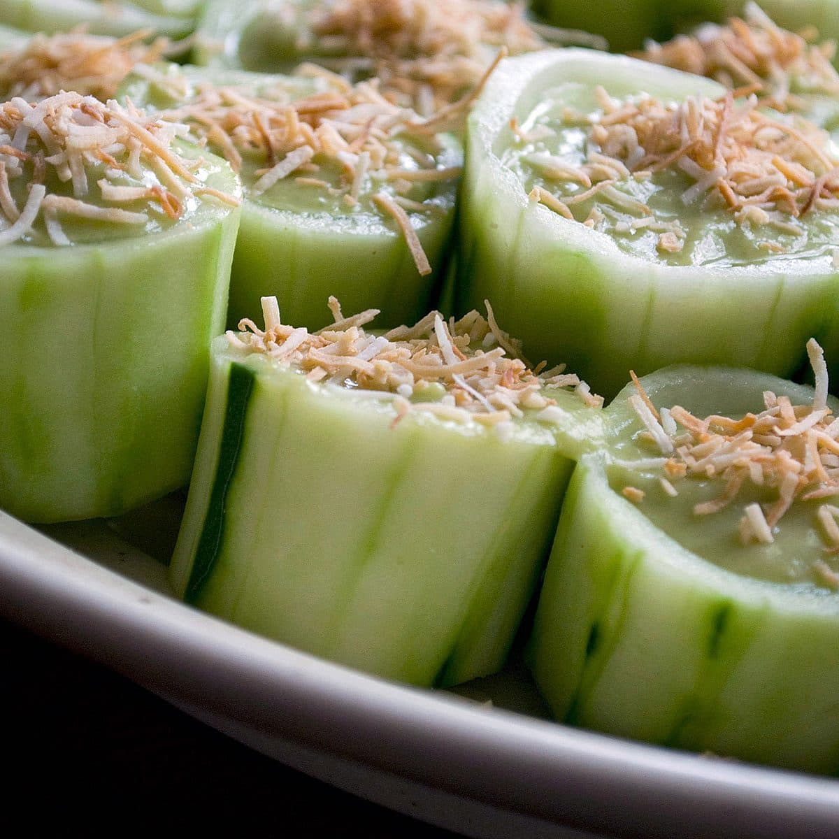 Close-up of sliced cucumbers filled with a creamy avocado mixture, topped with shredded cheese and arranged on a white plate. The cucumber slices are hollowed out to hold the filling, creating a visually appealing and fresh appetizer.