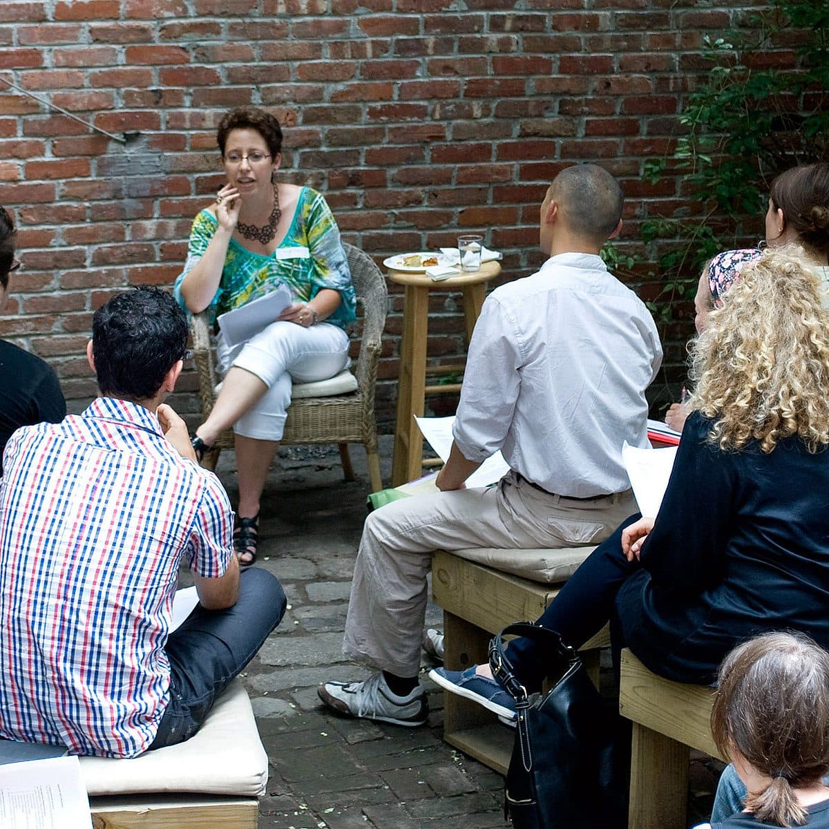 A group of people sits on benches and chairs in an outdoor space, attentively facing a woman who is speaking and holding notes. The setting has a relaxed, informal atmosphere with a brick wall in the background and greenery on one side.