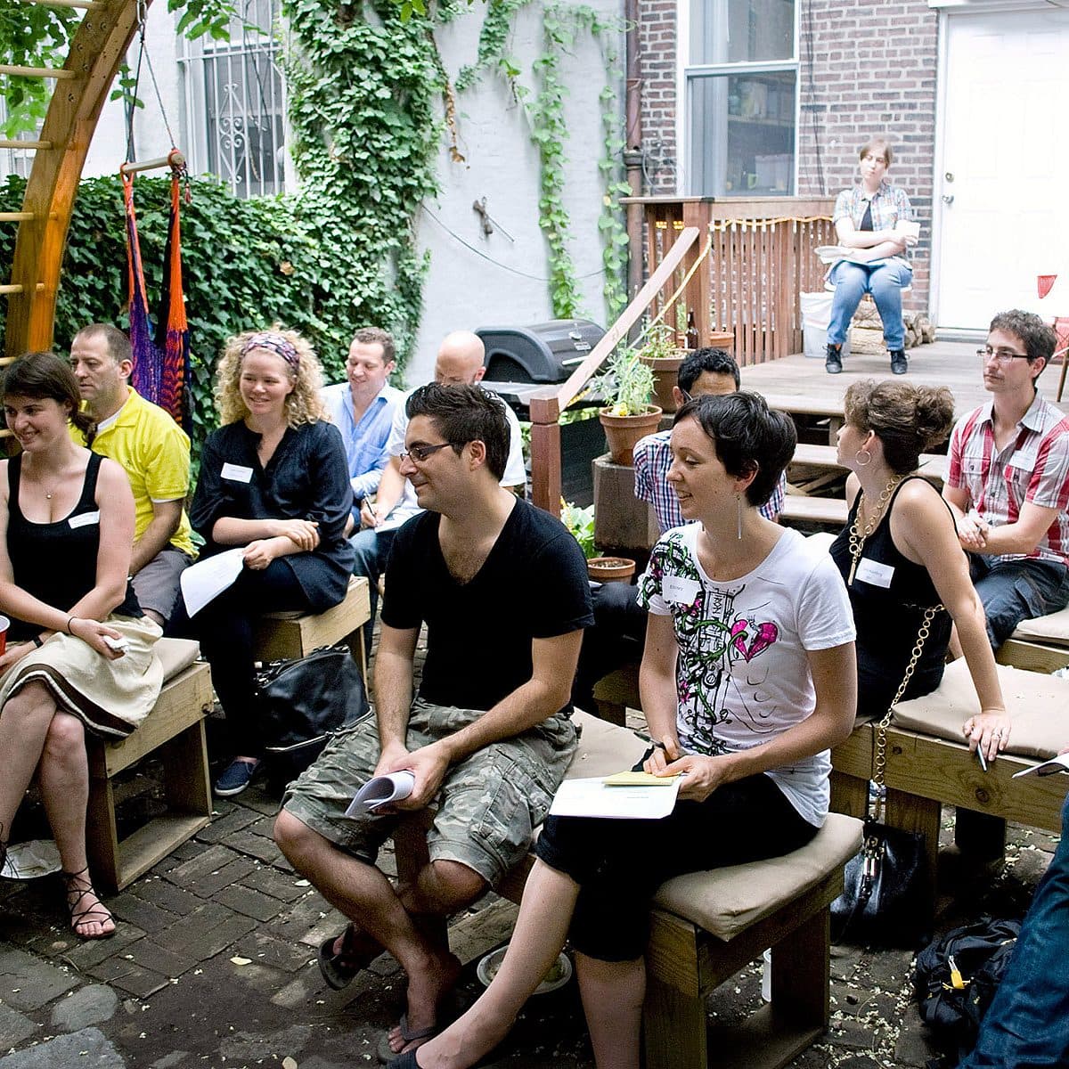 A diverse group of people sits attentively in an outdoor space with wooden benches and a brick wall covered in ivy. They are engaged in a meeting or workshop, with some taking notes. The setting appears casual, with some people holding notebooks and wearing relaxed attire.