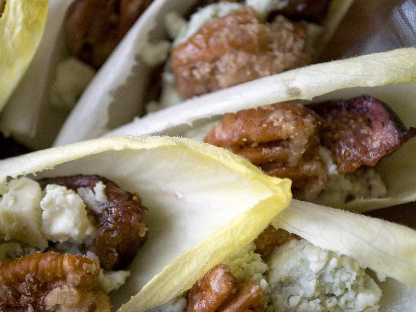 Close-up of endive leaves stuffed with walnuts, blue cheese crumbles, and caramelized ingredients. The colors are vibrant, with the light yellow endive contrasting against the darker brown nuts and creamy blue cheese.