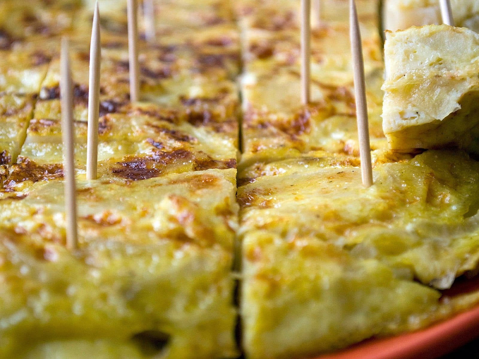 Close-up of sliced Spanish tortilla (potato omelet) pieces arranged on a plate, each with a toothpick inserted for easy picking. The tortilla is golden and slightly browned on the edges.