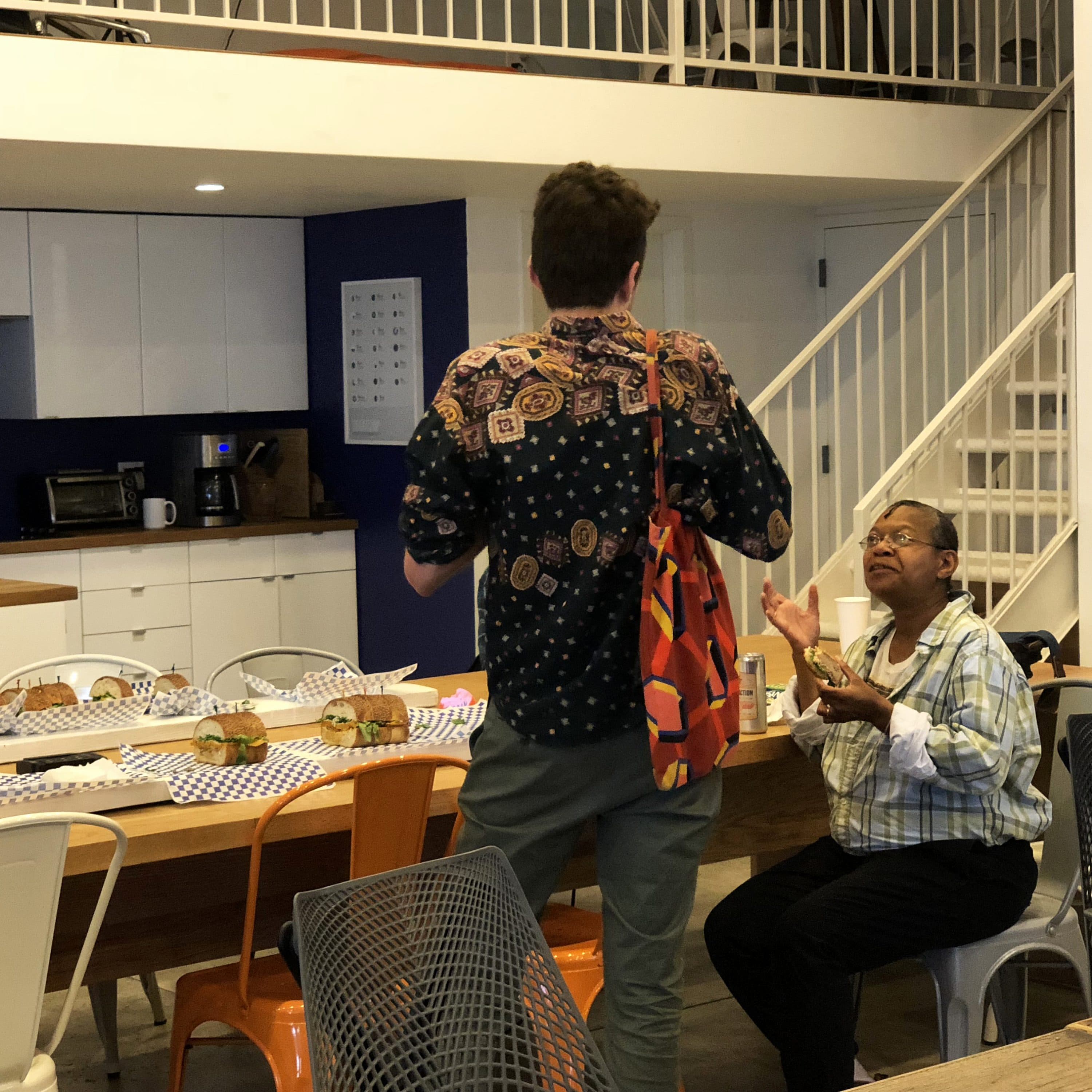 A man stands talking to a seated woman in a casual office break area. There is a long table nearby with sandwiches and drinks. The room features modern decor with white cabinets, a staircase, and people engaging in conversation.