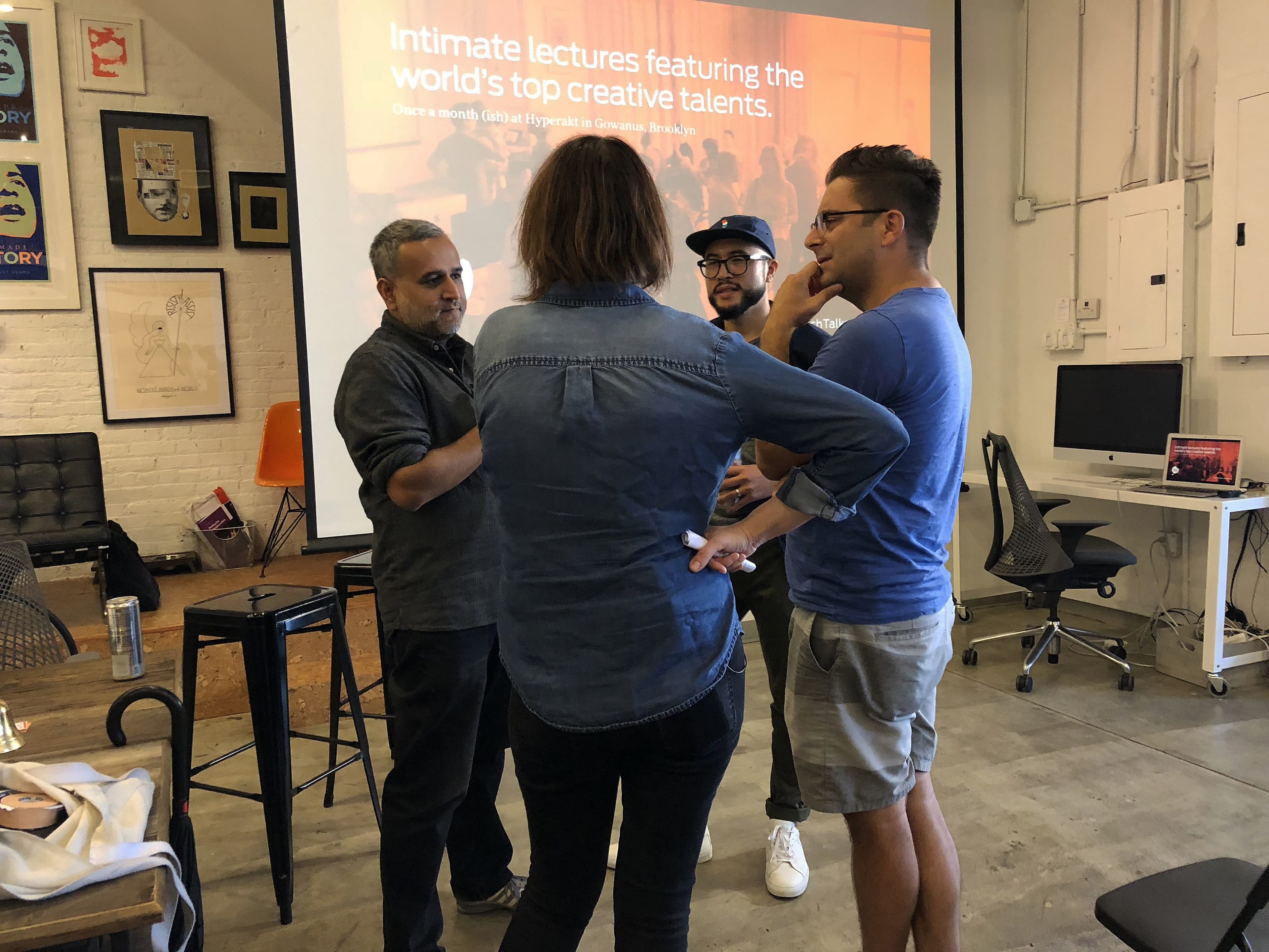 A group of four people stands in a creative workspace having a discussion. A projected screen behind displays text about lectures with top creative talents. The room has a whiteboard, framed artwork on brick walls, a table, and chairs.