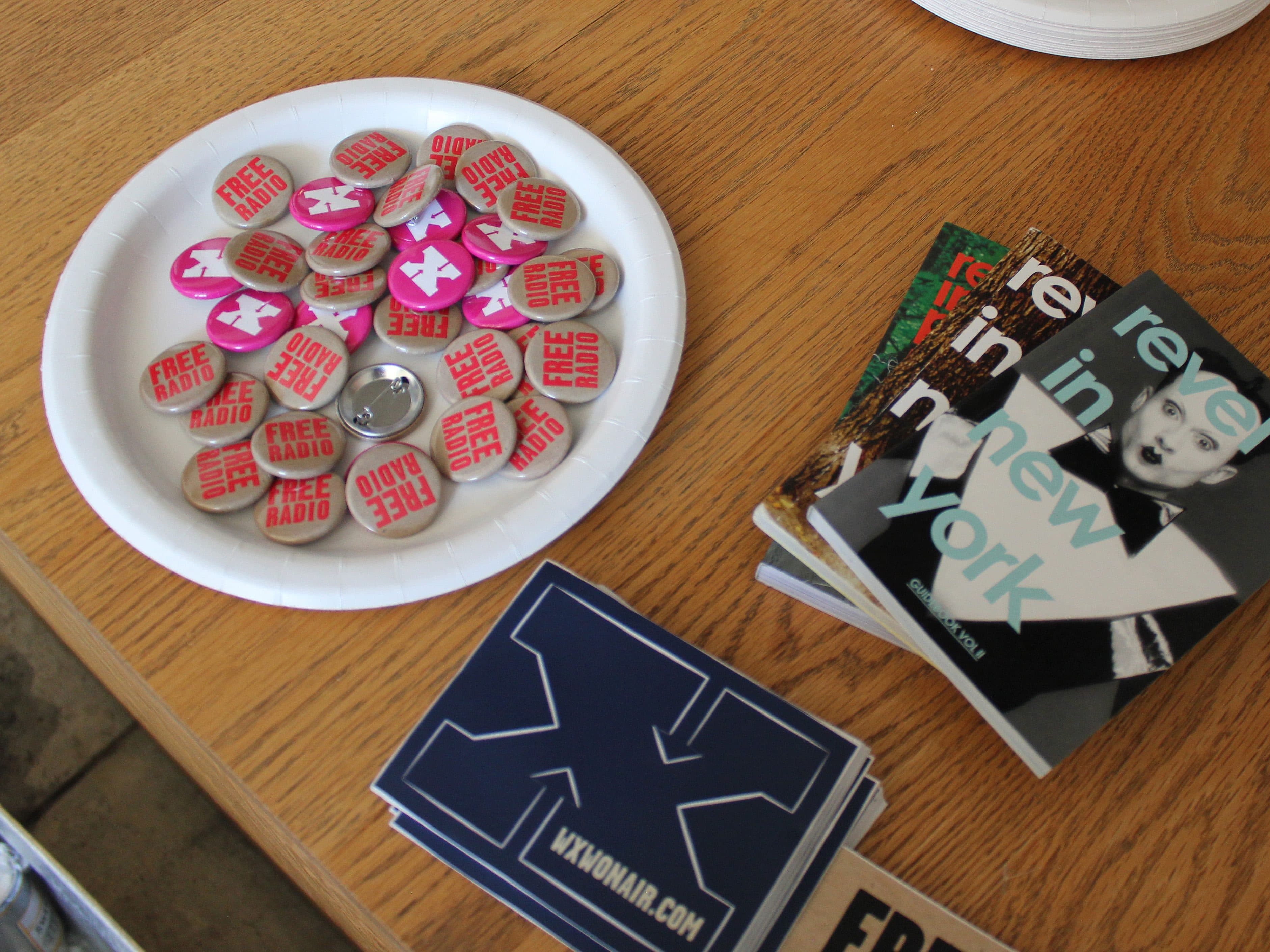 A table contains a paper plate filled with pink and red "Free Radio" buttons, a stack of stickers with a large blue letter "X," and three "revel in New York" booklets featuring a black and white photo of a person on the cover.