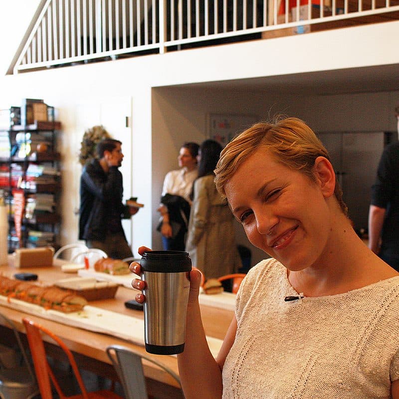 A smiling person with short hair holds a travel mug in a casual, indoor setting. Behind them, several people are gathered near a long table with food and drinks. Shelves with various items are visible along the wall, and a staircase leads to an upper level.