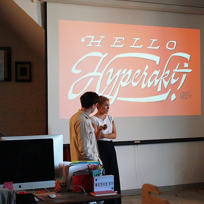 A man and woman stand in front of a large screen during a presentation. The screen displays a red slide with white cursive text that reads "Hello Hyperakt!" The room is an office setting with computers, framed art on the walls, and minimal decor.
