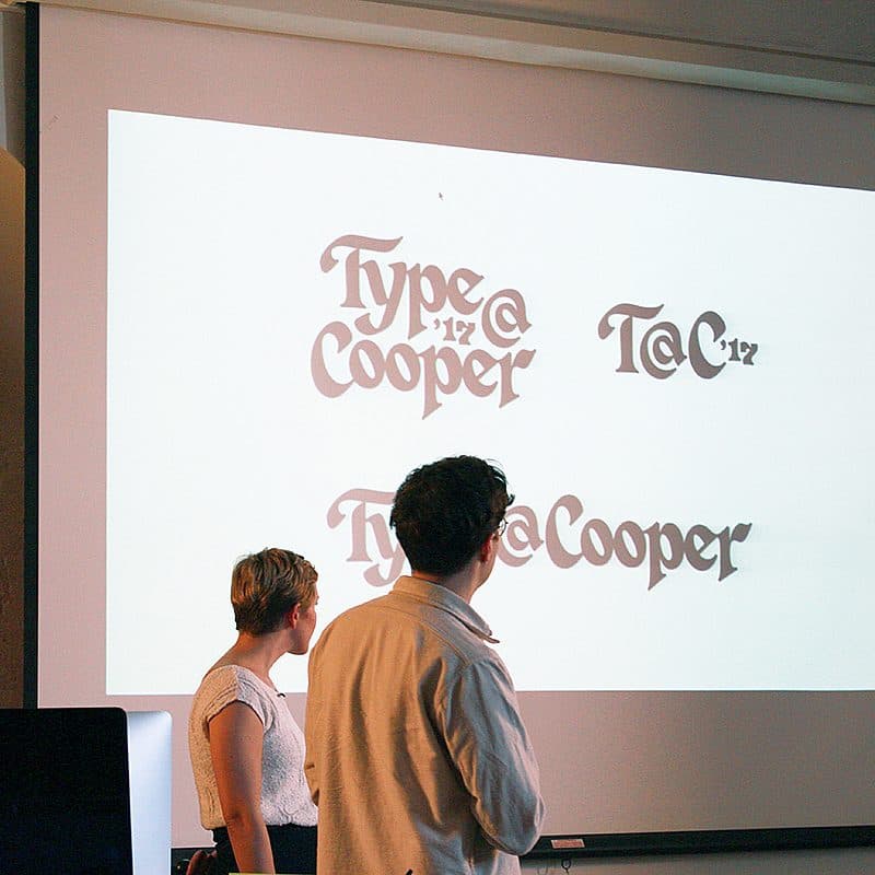Two people stand in front of a large projector screen displaying various stylized text samples reading "Type@Cooper." They appear to be discussing the typography designs. The room is dimly lit, with framed pictures on the wall in the background.