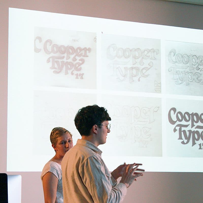 A man and woman stand in front of a large screen displaying six sketches of different 'Cooper Type' typography designs. The man is gesturing with his hands as he speaks, while the woman stands beside him, observing. The background is dimly lit.