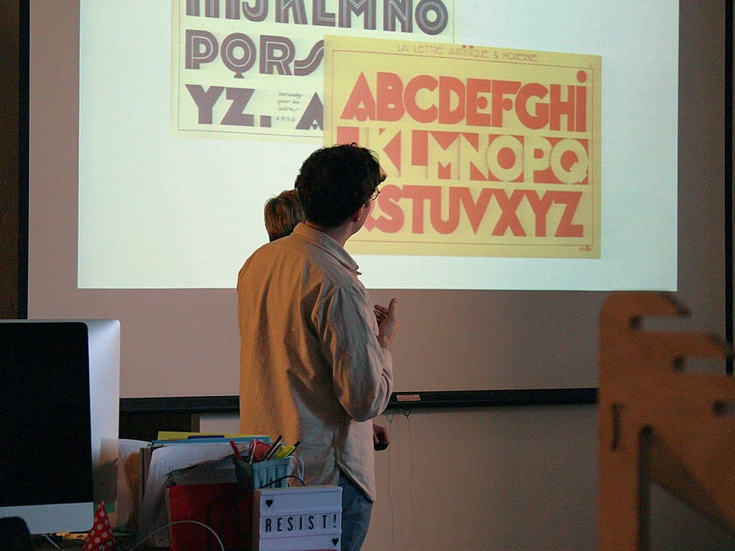 Two people standing in front of a projection screen displaying bold, vintage-style alphabet typography. The screen shows both uppercase and lowercase letters. The individuals appear to be discussing or presenting the content. Various office items are in the foreground.
