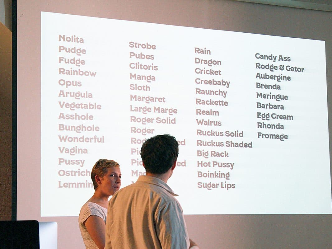 Two people stand in front of a projected screen displaying a list of names or words. The list includes terms such as "Nolita," "Pudge," "Clitoris," "Dragon," "Rain," "Candy Ass," and "Fromage." The room appears to be dimly lit with the focus on the screen.