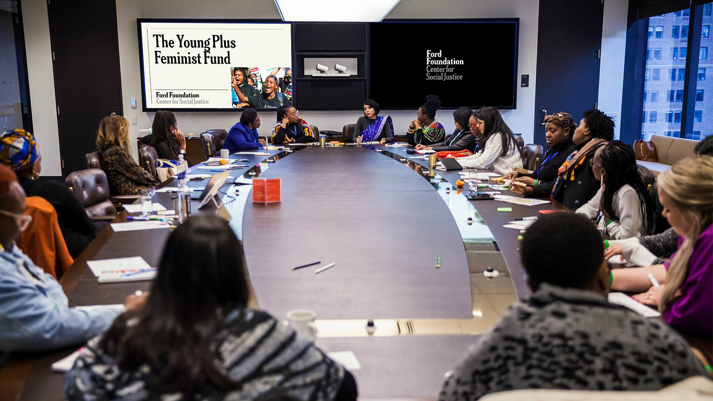 Group of people sitting around a large oval conference room table with screens on the wall containing a Ford Foundation branded presentation