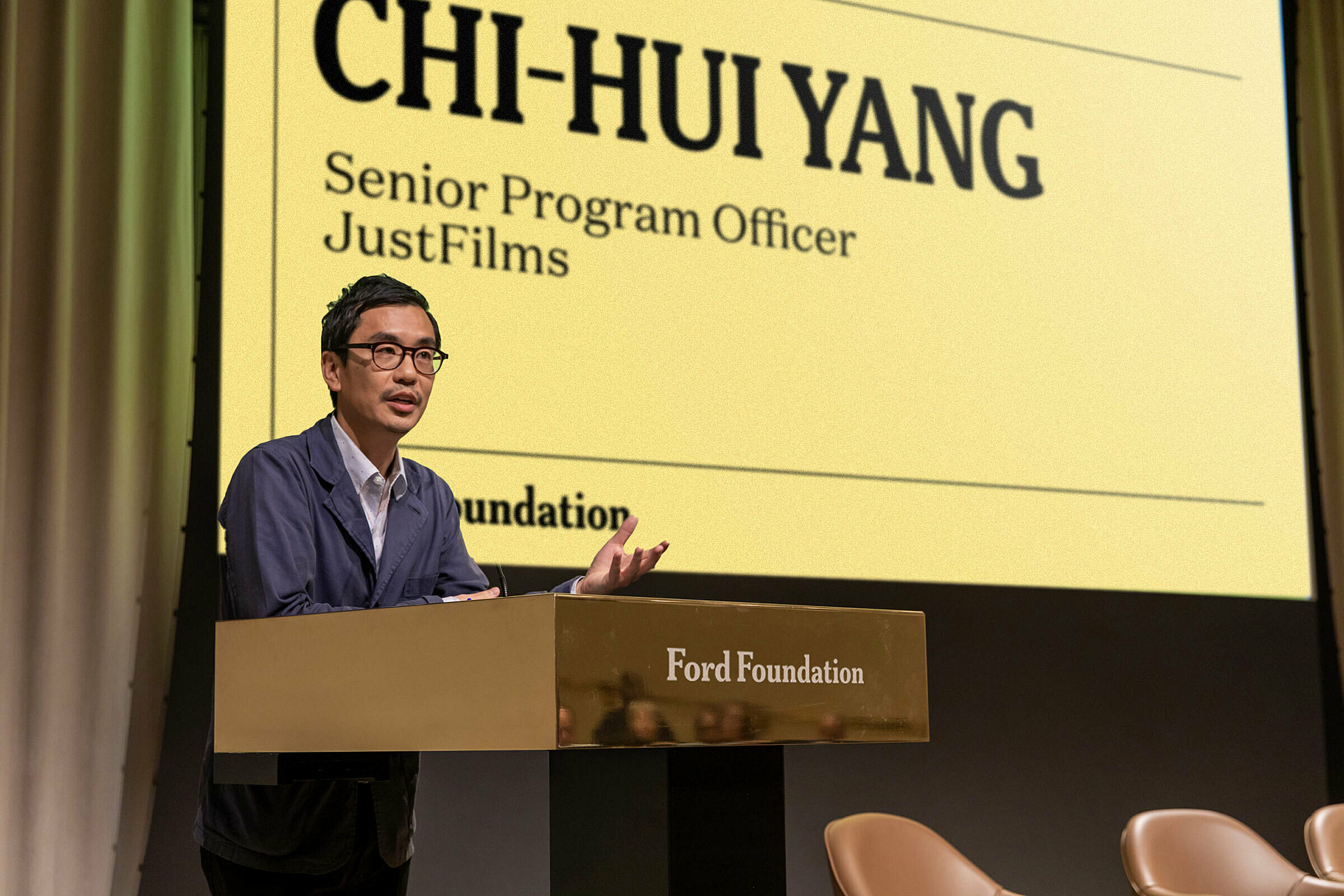 Chi-Hui Yang presenting on behind a Ford branded podium with yellow Ford branded presentation slide projected behind him